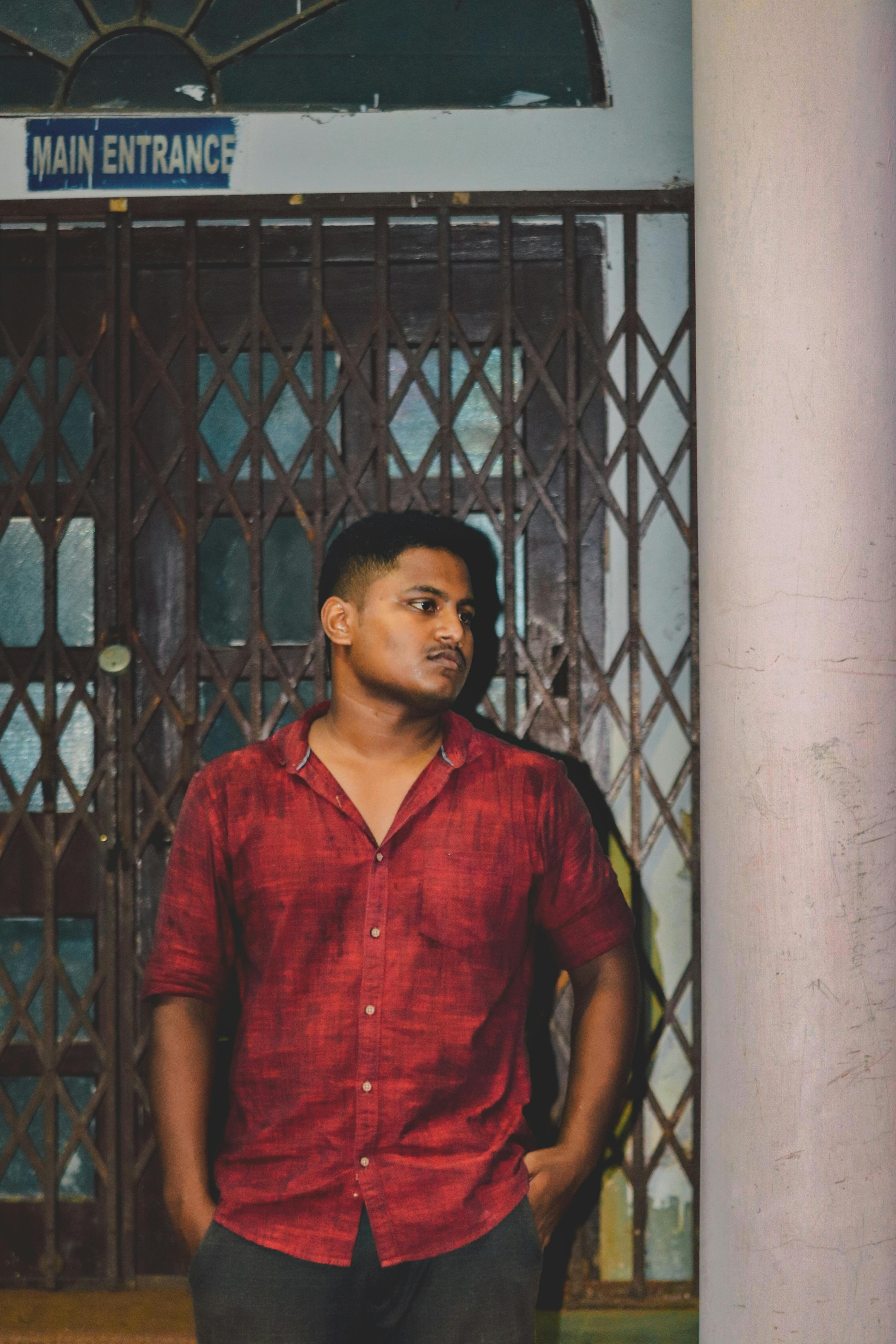Young man in red shirt leaning against pillar near metal entrance gate at night.