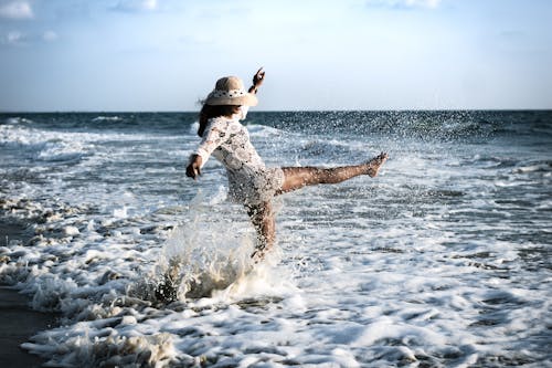 Woman in a Crochet Beach Dress and Sun Hat Kicking Sea Wave
