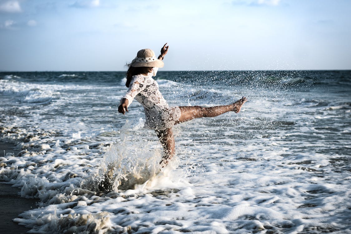 Woman in a Crochet Beach Dress and Sun Hat Kicking Sea Wave