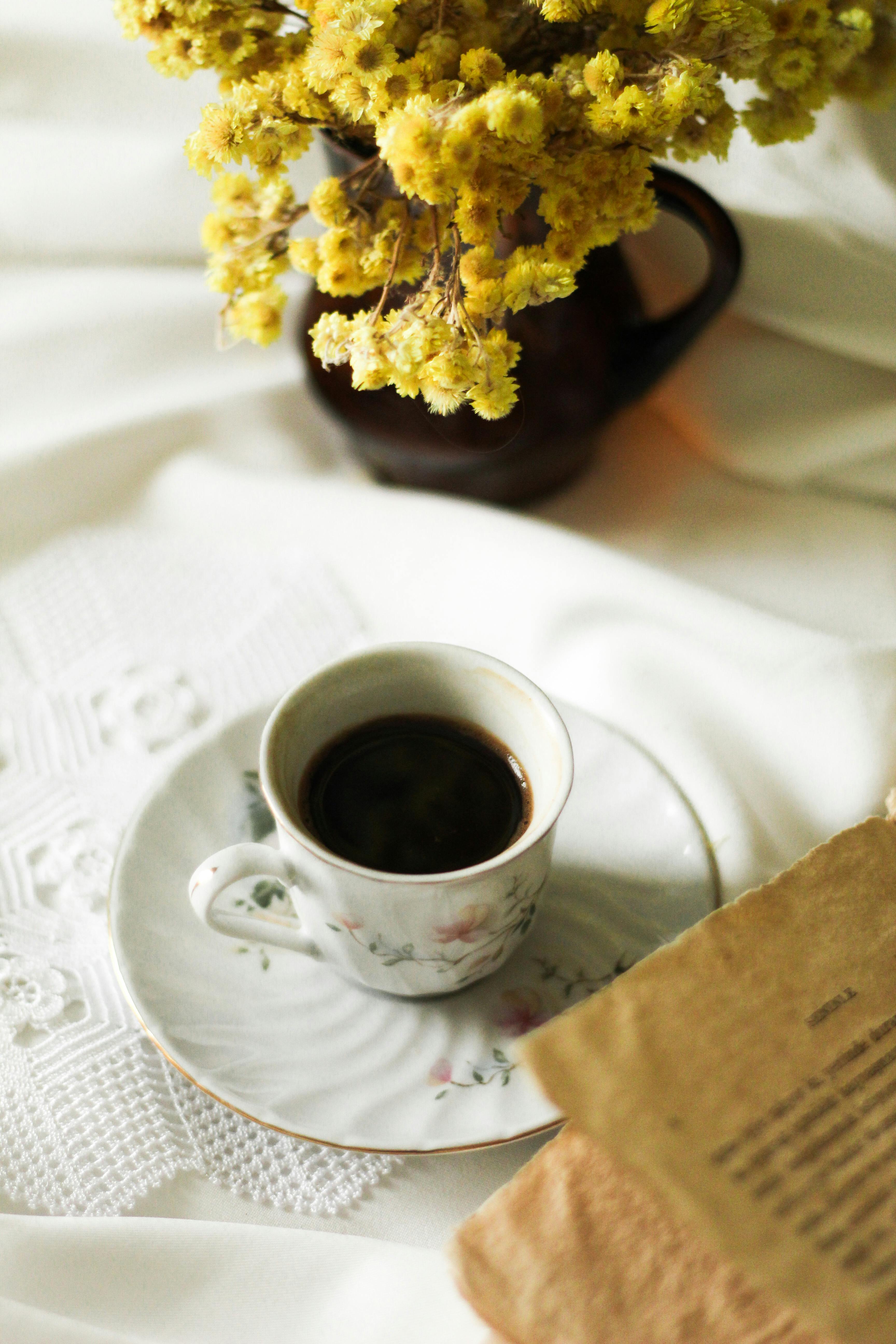 A beautiful still life of coffee in vintage porcelain, a book, and yellow flowers on a white tablecloth.