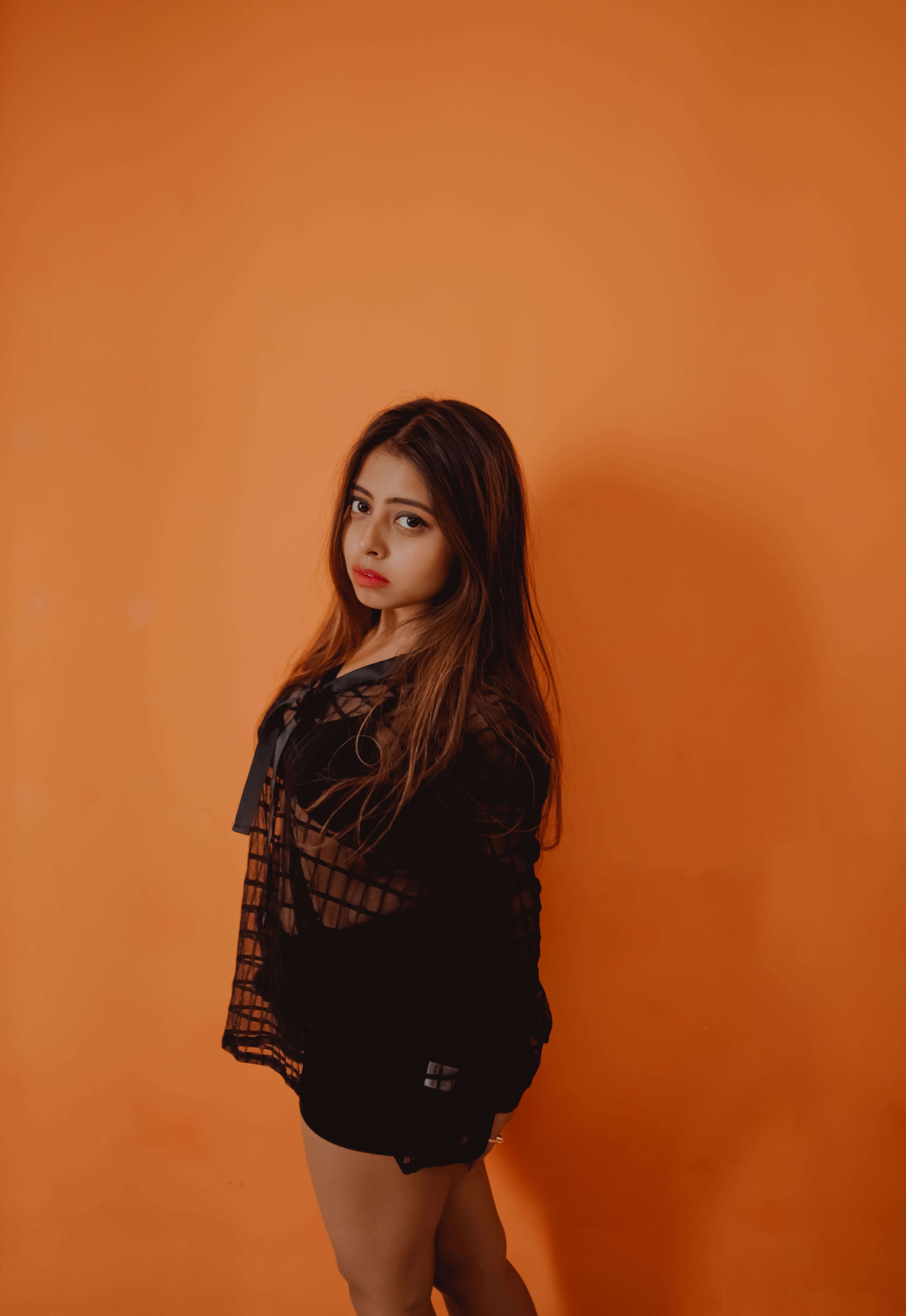 A fashionable young woman in a black outfit poses confidently against a bold orange background.