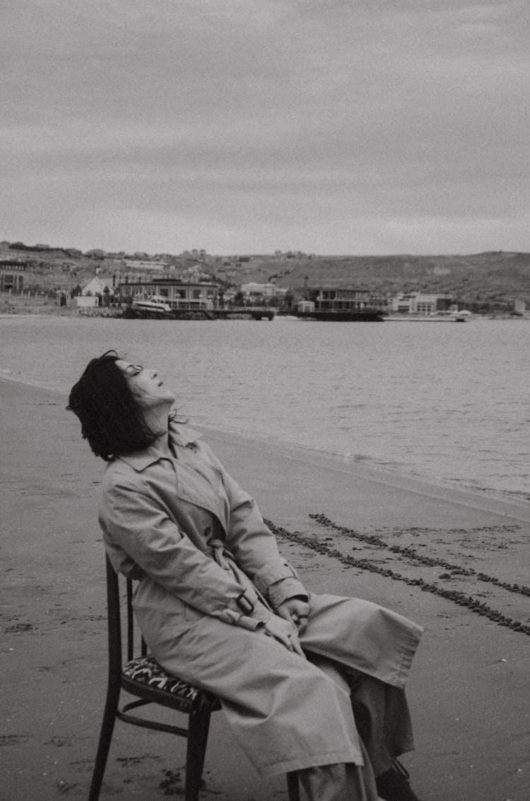 Woman In Trench Coat Sitting On Chair At Beach