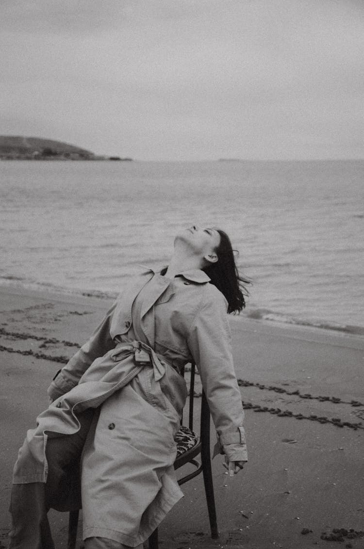 Woman In A Trench Coat Sitting On A Chair On A Beach 