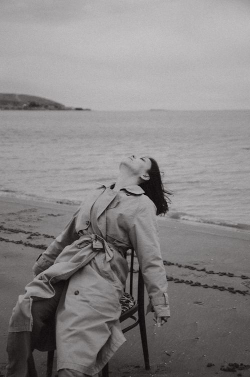 Free Black and white photo of a woman in a trench coat sitting on a chair at a beach in Baku. Stock Photo