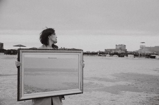 A woman standing on a Baku beach holding a framed reflection, captured in black and white.