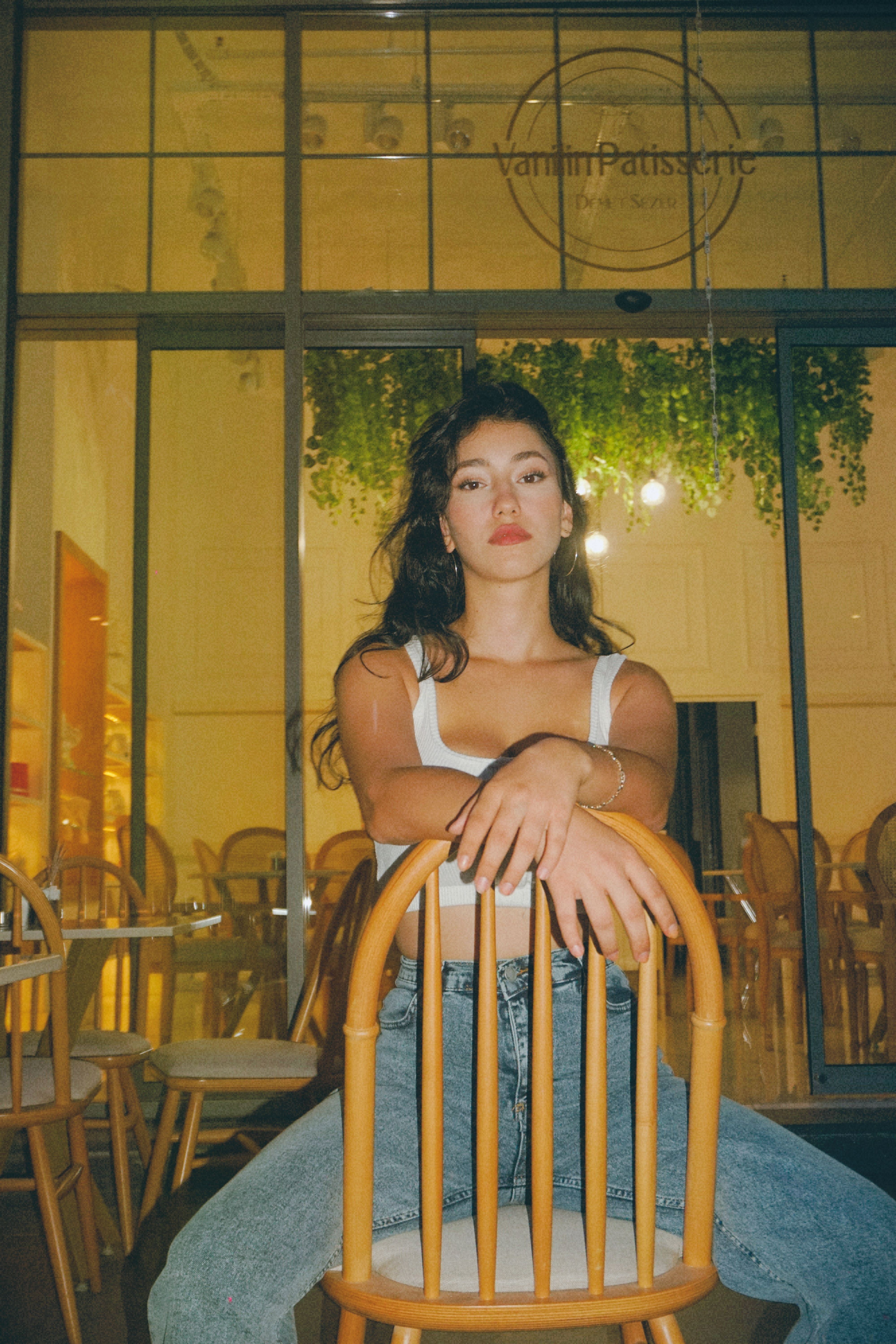 A fashionably dressed woman poses on a chair in front of an urban patisserie at night, showcasing street style.