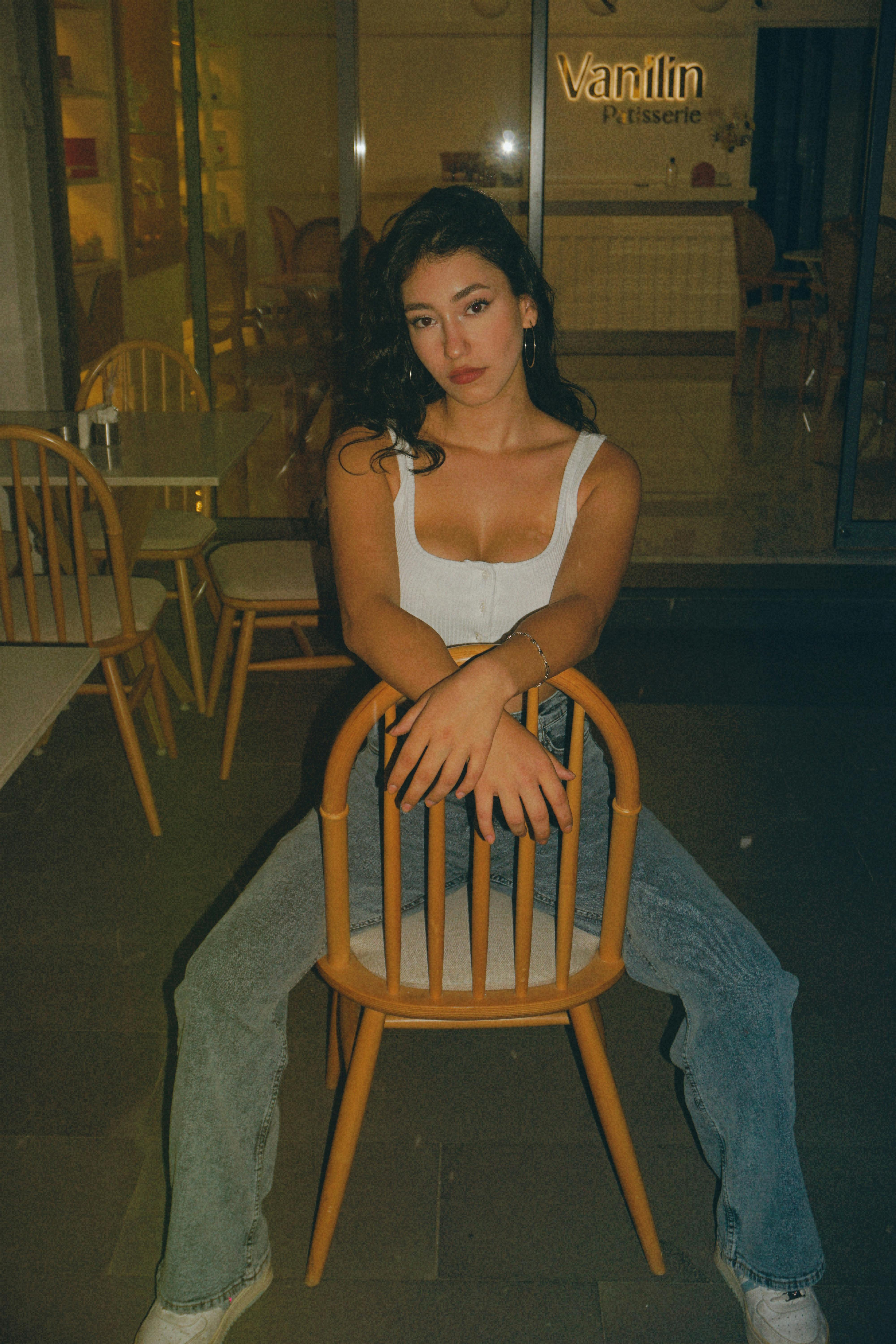 Stylish young woman in a tank top and jeans posing on a chair indoors at night.