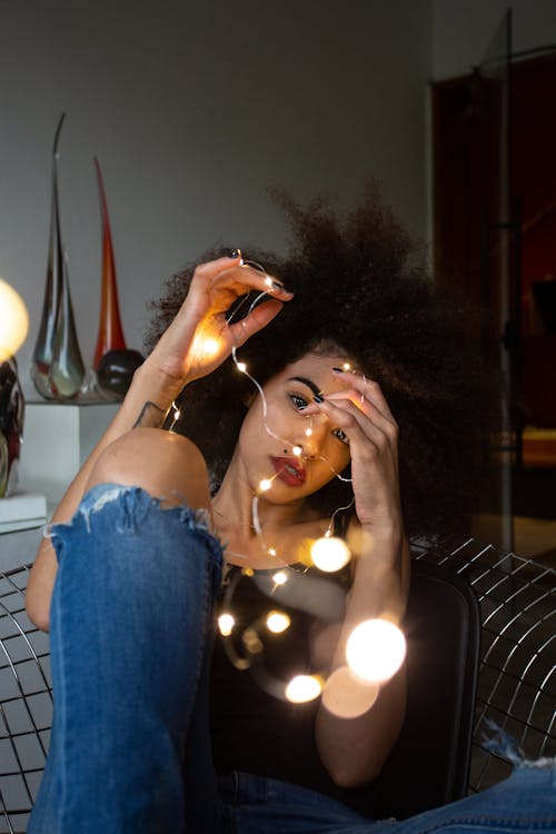 Woman in Black Tank Top Sitting on Chair and Holding Lighted String Lights