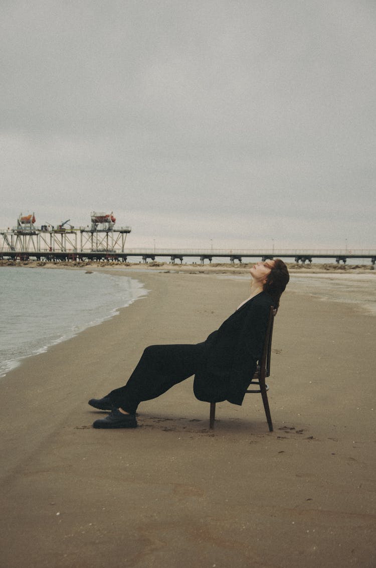 Woman In Black Suit Sitting On Chair At Beach Against Pier