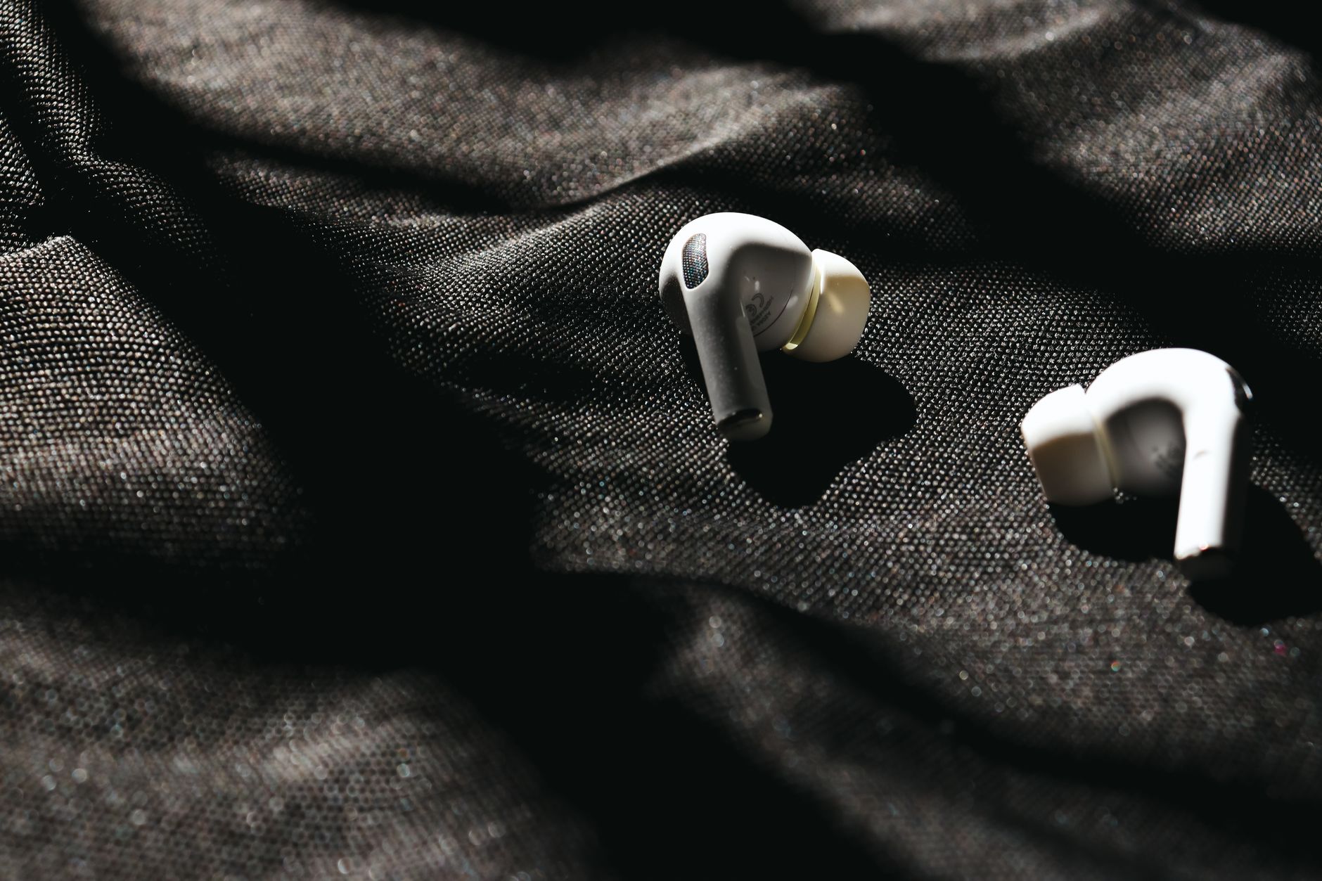 Close-up of white wireless earbuds on black fabric with dramatic sunlight and shadows.