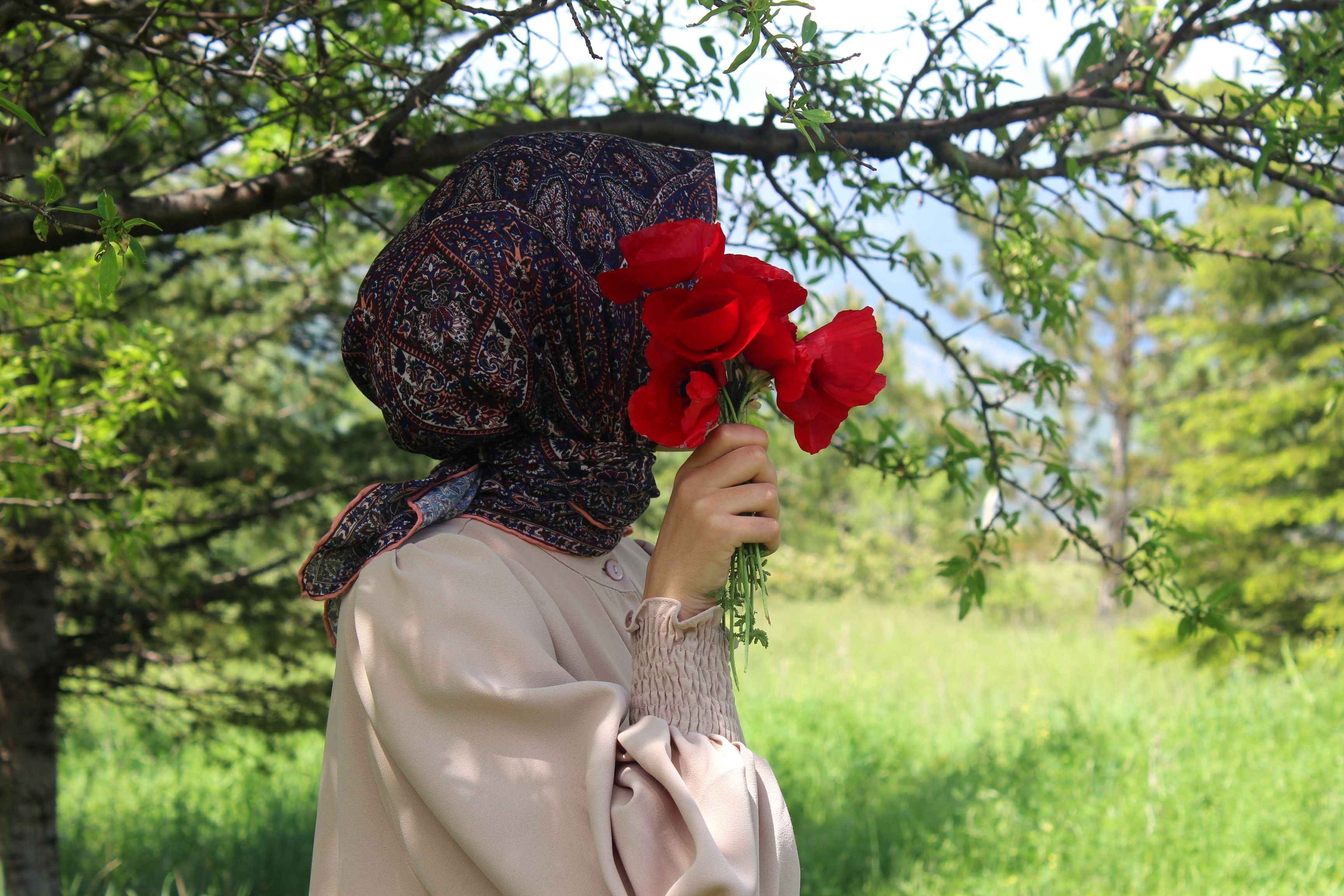 A woman in a headscarf holds bright red flowers in a lush green summer field.