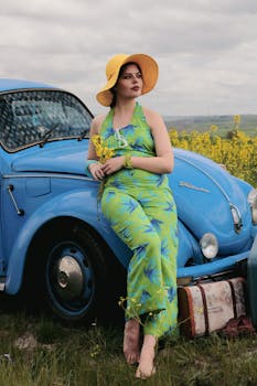 Woman in vintage dress leaning on blue car with suitcases in rural field.