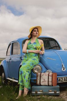 Stylish woman in a summer dress leaning on a vintage car in the countryside.