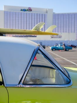 Close-up of a vintage car with a surfboard on top in a parking lot, sunny day.