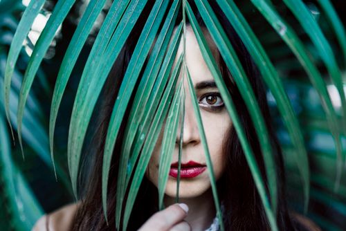 Free Portrait of a woman with vivid makeup, partially concealed by green palm leaves, creating a mysterious, natural look. Stock Photo