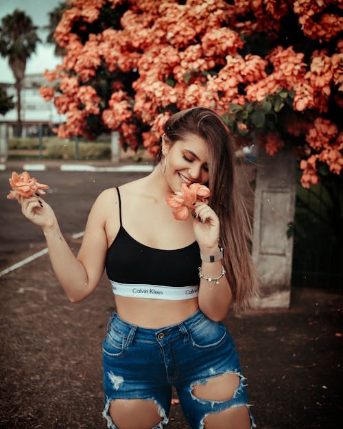 Free Woman in Black Tank Top and Blue Denim Shorts Standing Near Red Flowers Stock Photo