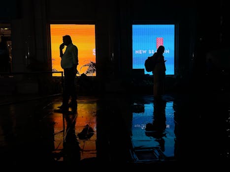 Two silhouettes in a dark corridor with vibrant billboard reflections creating a cinematic urban mood.