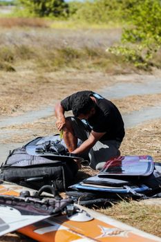 Focused man repairing kiteboard by the beach with scattered equipment in sunny weather.