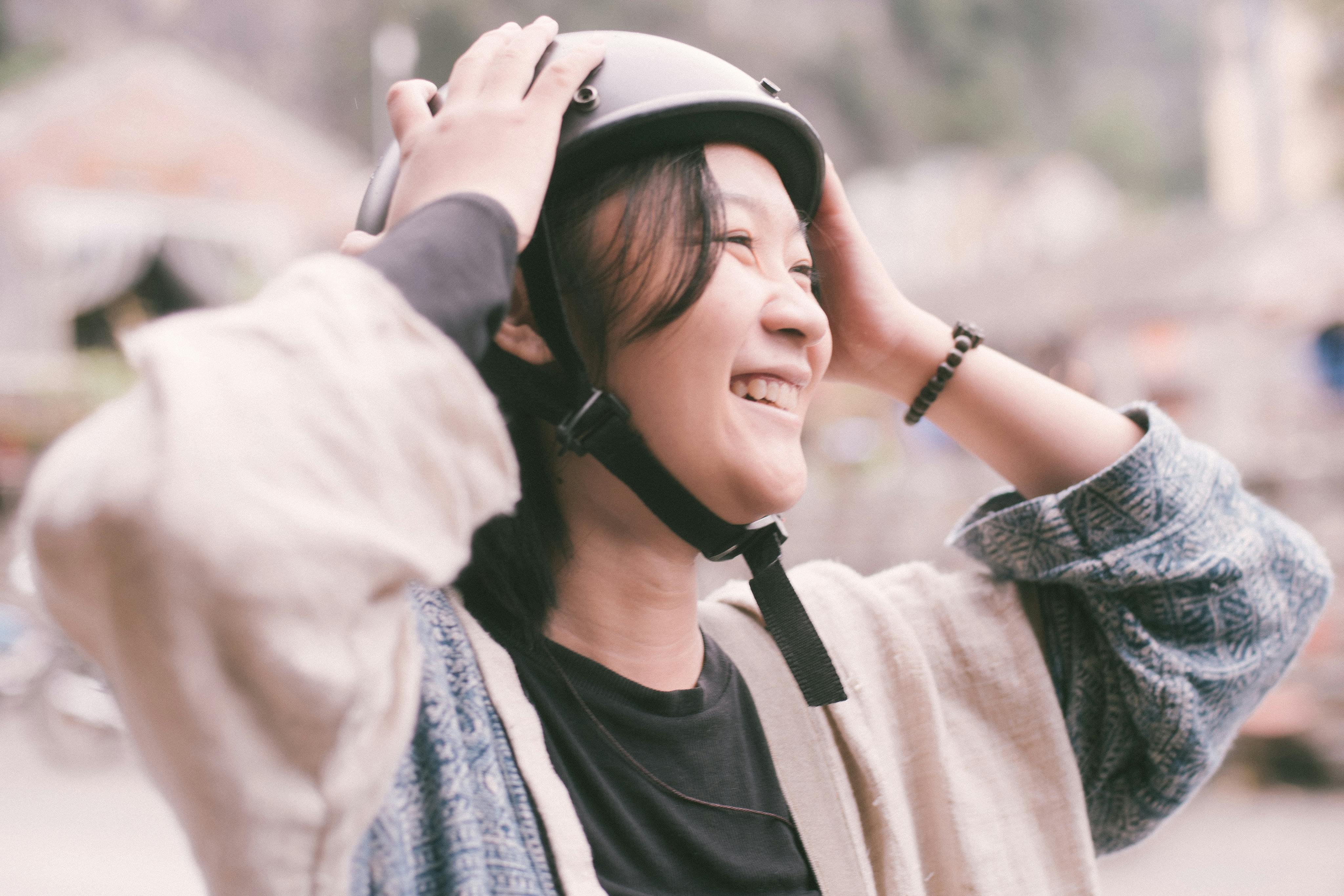 A cheerful young woman outdoors, adjusting her helmet with a big smile.