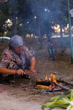 A woman crouches by a bonfire roasting corn in an outdoor setting with smoke rising.