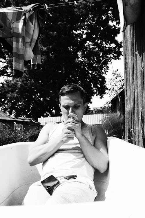a guy smoking a cigarette lying in a white bathtub outside a rustic house