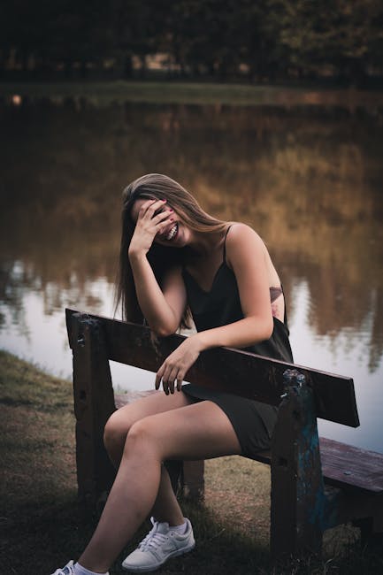 Photo of Laughing Woman in Black Dress Sitting on Brown Wooden Bench by a Lake