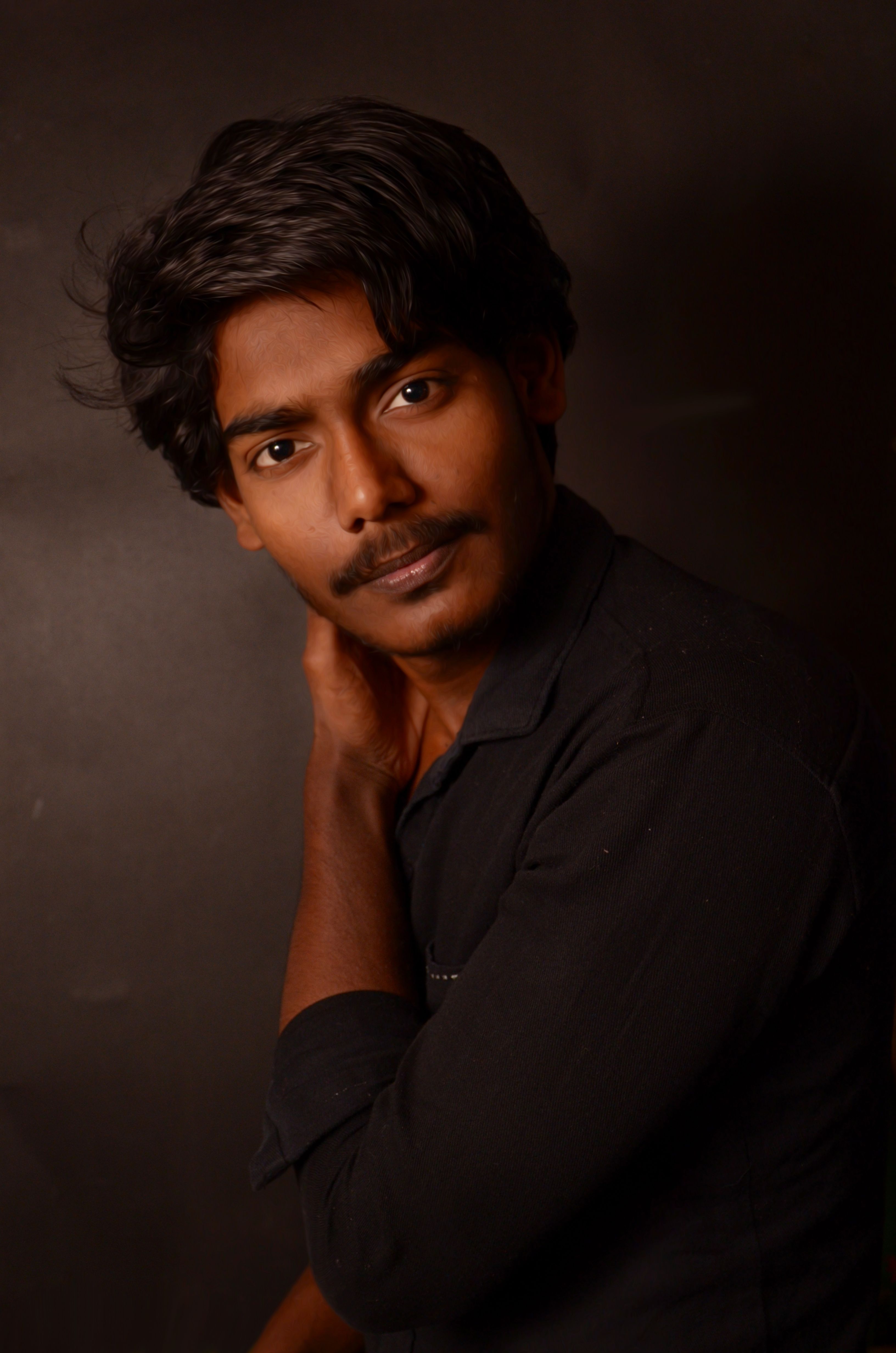 Intimate portrait of a young South Asian man in a black shirt posing against a dark background.