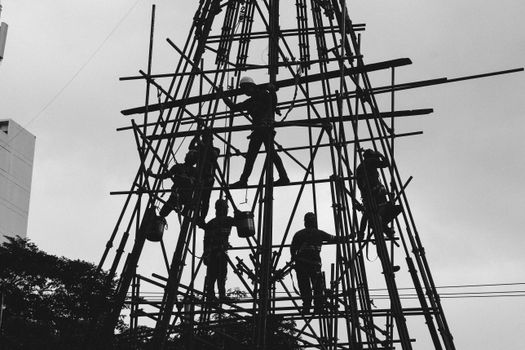 Silhouetted workers construct scaffolding against the urban skyline in black and white.