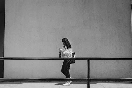 Black and white photo of a woman walking with a smartphone against a large concrete wall.
