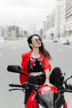 Stylish woman in red jacket and sunglasses sitting on a motorcycle in urban Baku.