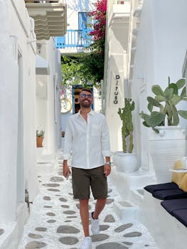 Young man strolling in a sunlit alley of Mykonos, Greece, wearing khaki shorts and white shirt.