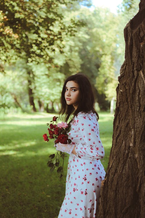 Portrait of a Pretty Brunette Leaning on a Tree with Red Flowers in Hands
