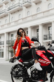 Stylish woman poses on motorcycle in urban Baku street wearing red jacket and sunglasses.