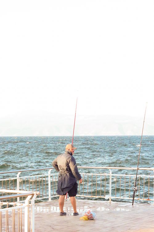 Free A fisherman in a raincoat stands at a pier with fishing rods on a bright day. Stock Photo