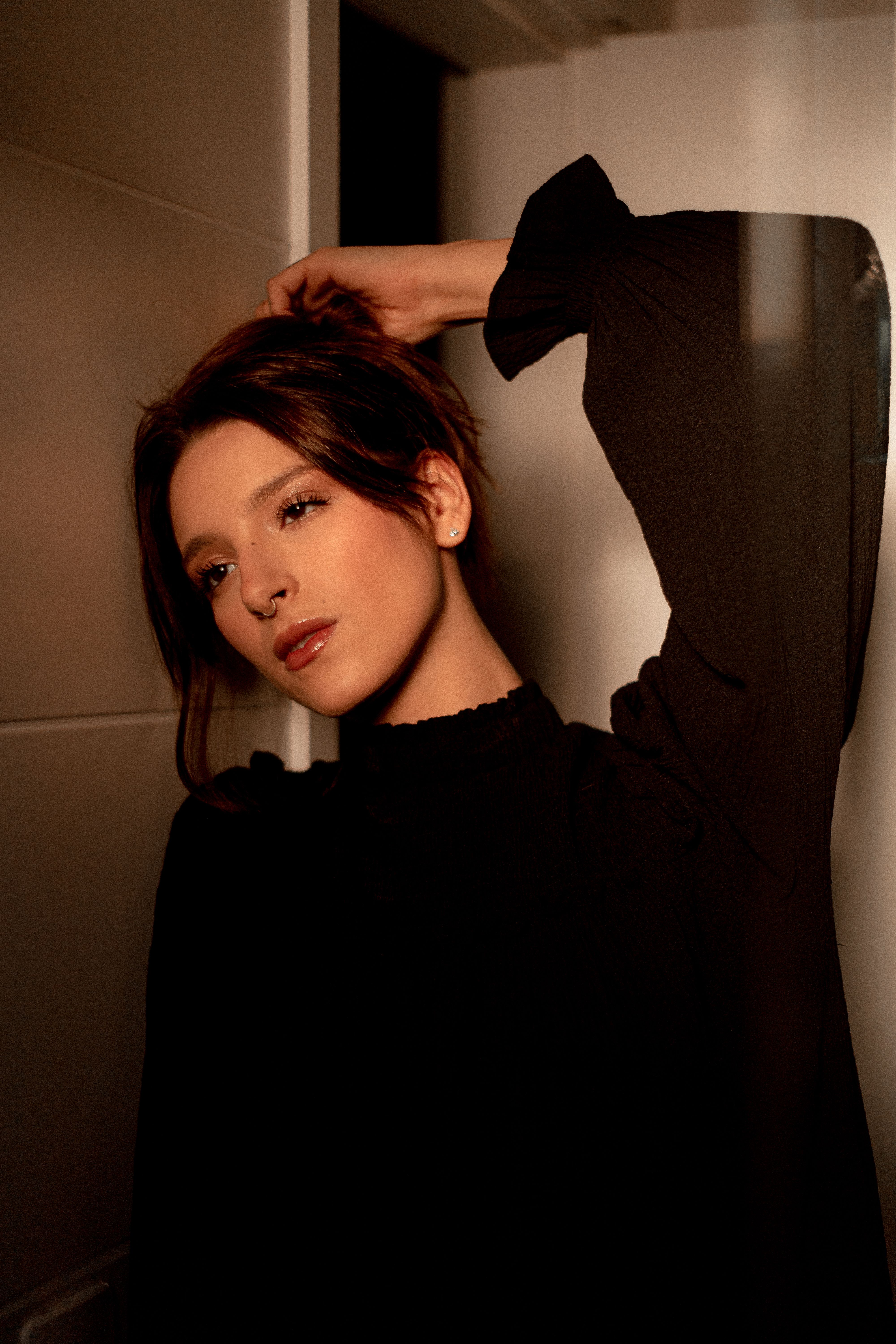 A woman in black attire gracefully poses, capturing warm, soft lighting indoors for a fashion shoot.