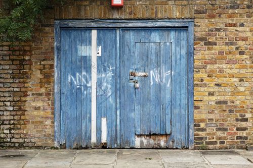 Free A rustic blue wooden garage door set against a weathered brick wall, featuring graffiti. Stock Photo