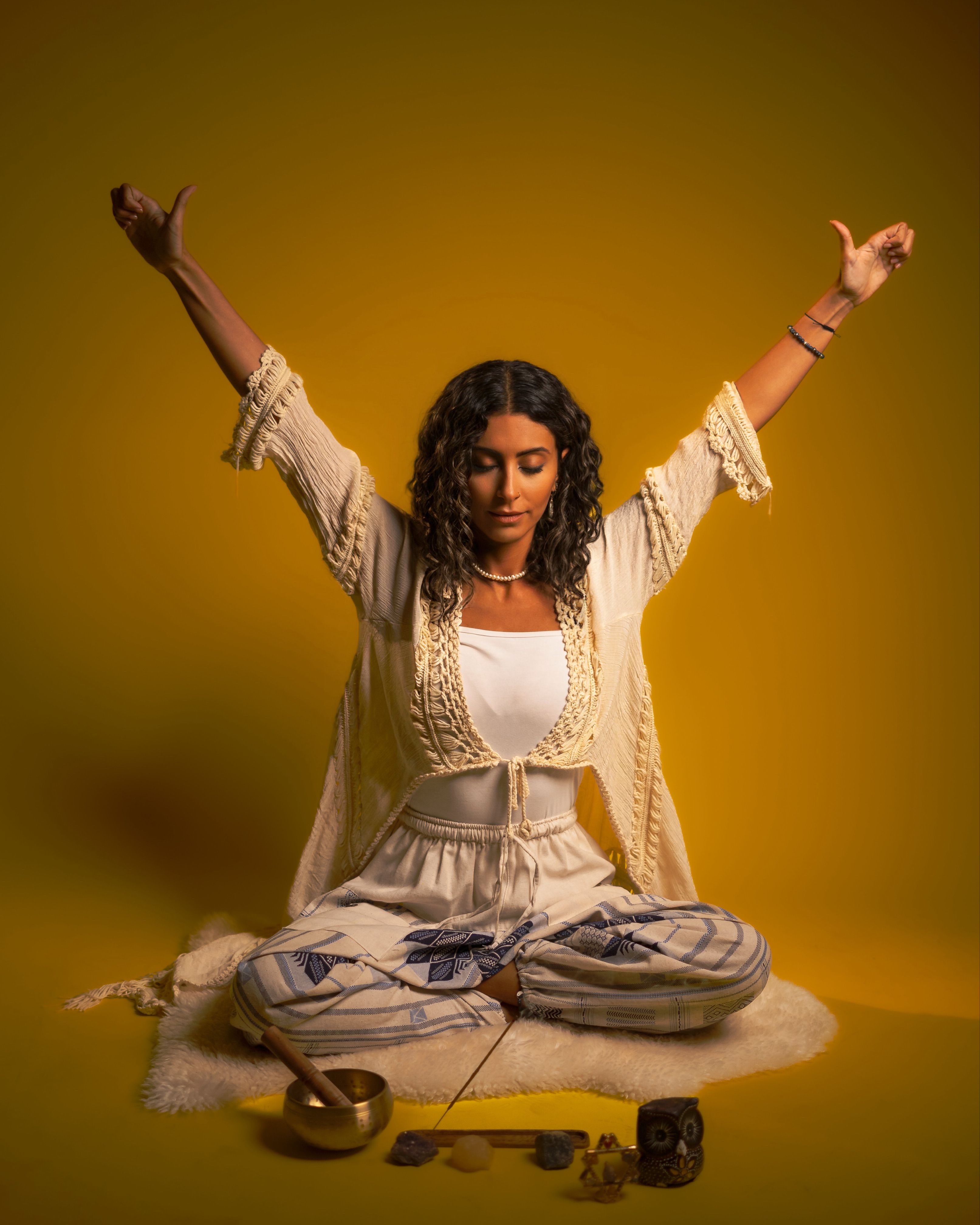 Woman meditating with raised arms and a singing bowl against a warm yellow background.