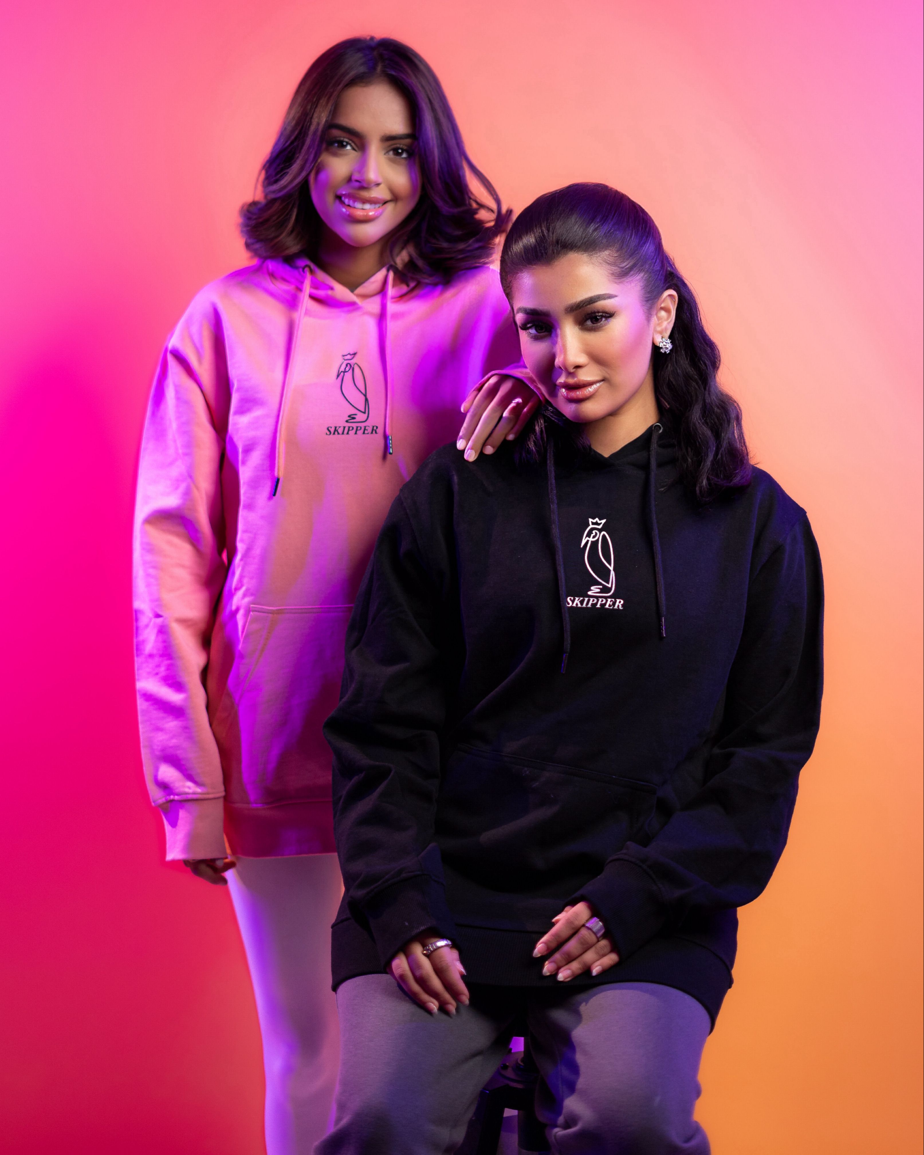 Two women in vibrant hoodies pose against a colorful backdrop.