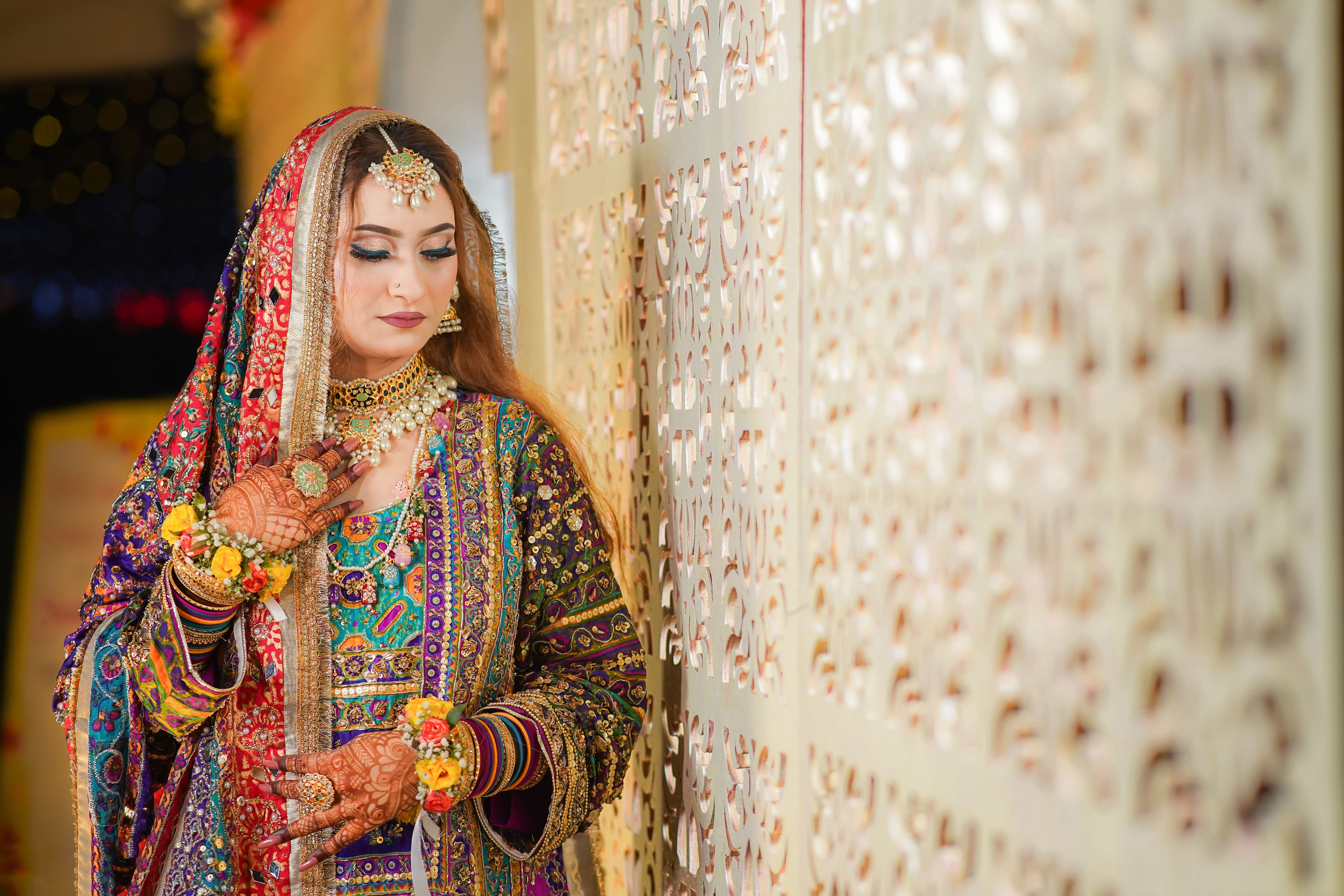 South Asian bride wearing colorful traditional attire with intricate patterns and jewellery, posing in a decorative setting.