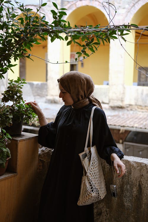 A woman in a black dress and headscarf is standing near a plant