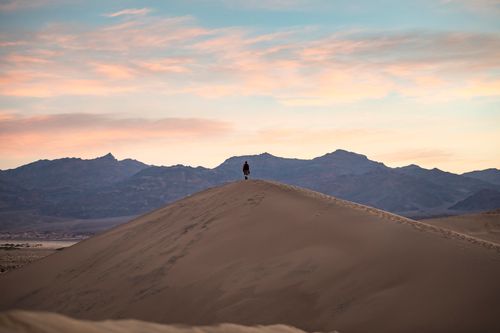 Free A lone hiker stands atop a sand dune with a dramatic dusk sky and mountains in the background. Stock Photo