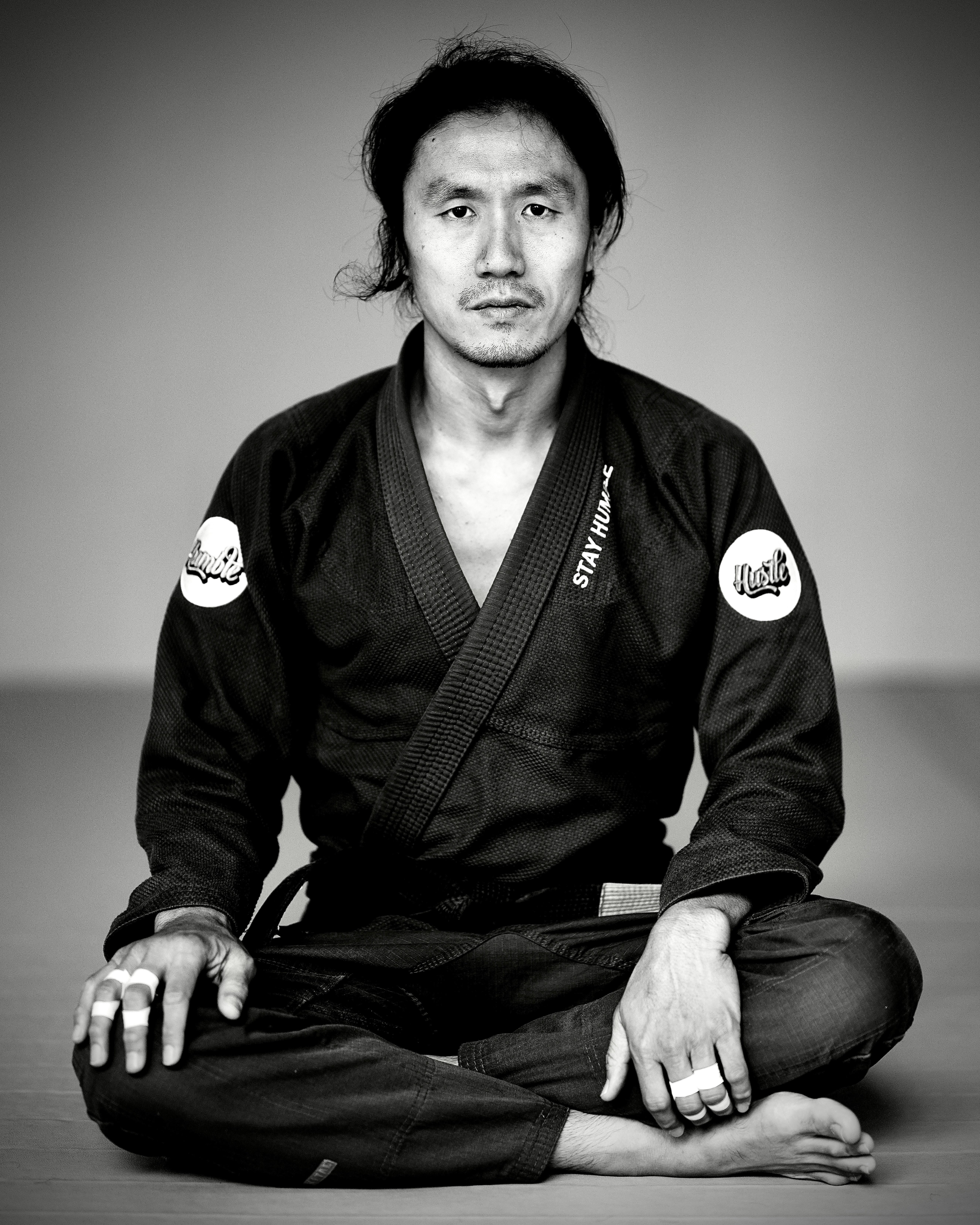 Monochrome portrait of a martial artist in a kimono, sitting cross-legged in a studio.