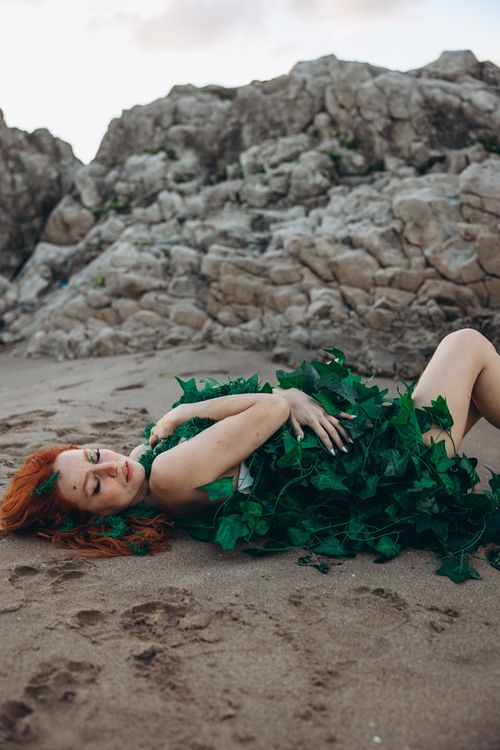 Free Woman in green costume with red hair lying on a rocky beach, surrounded by nature. Stock Photo