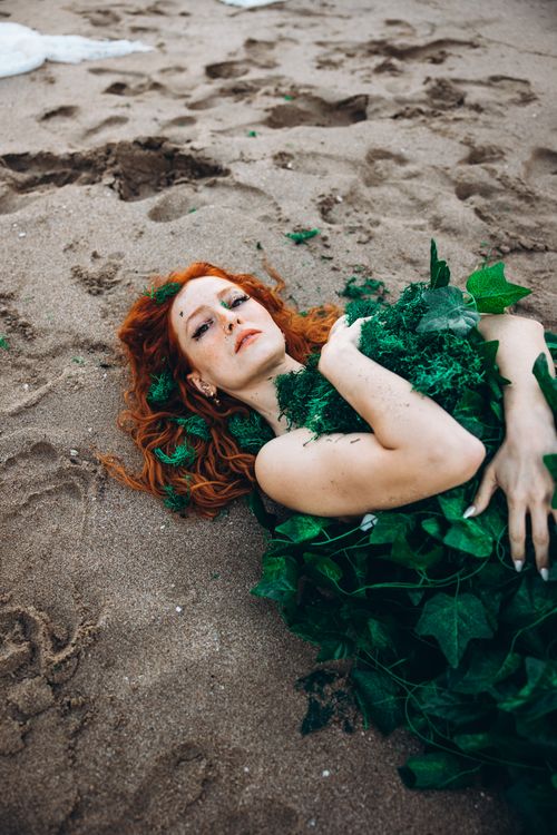 Free Red-haired woman lying on sandy beach covered in green ivy leaves. Stock Photo