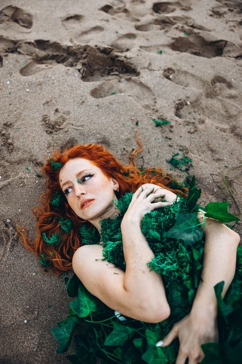 Free Red-haired woman with greenery artfully draped, lying on sandy beach. Stock Photo