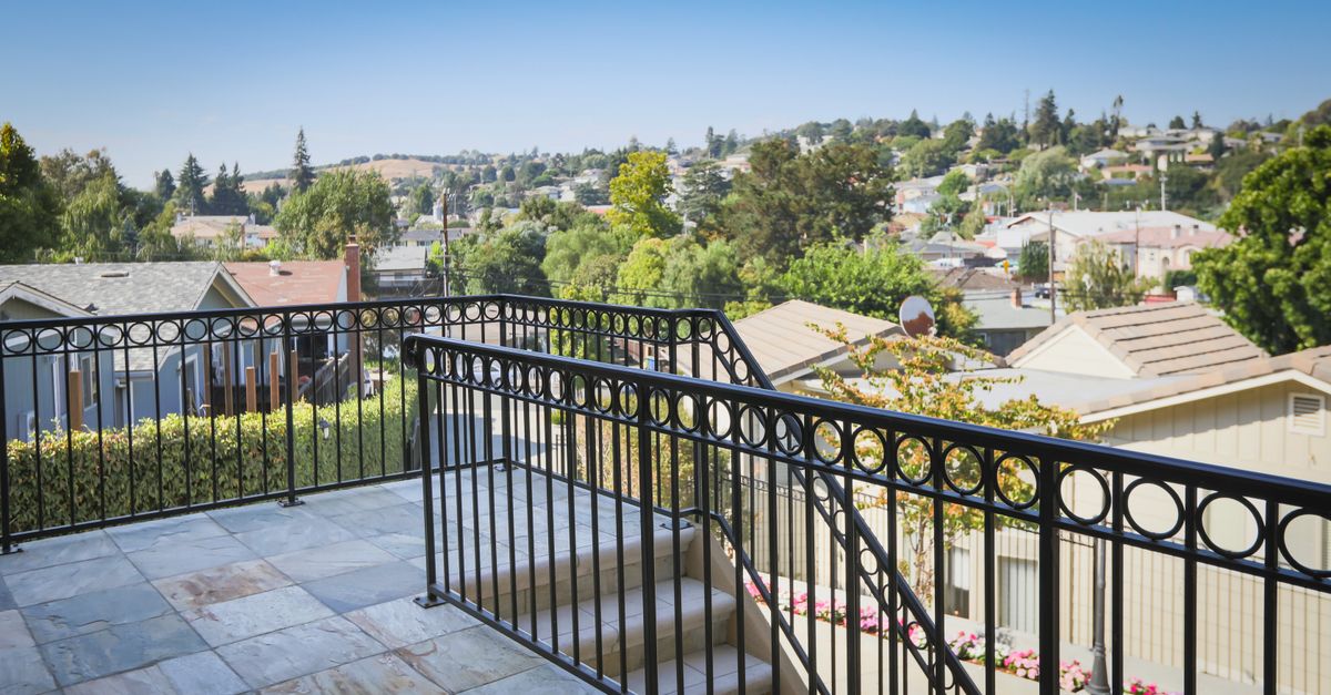 A sunny suburban neighborhood view from a balcony with black metal railing and lush greenery.