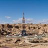 A towering oil drilling rig operates in the center of a rugged, arid desert landscape.