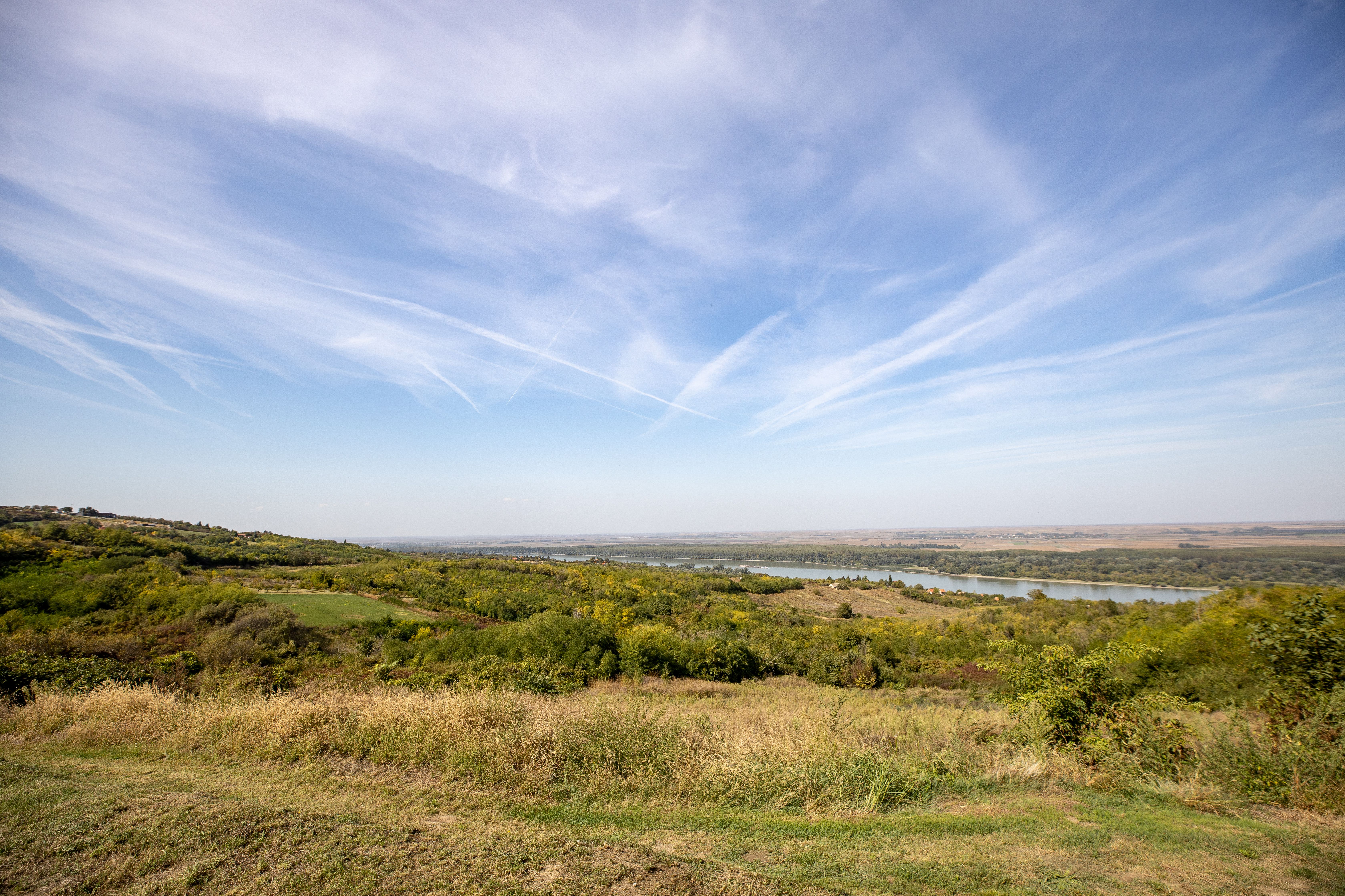 A vast countryside landscape featuring rolling hills, green fields, and a river under a bright blue sky with clouds.
