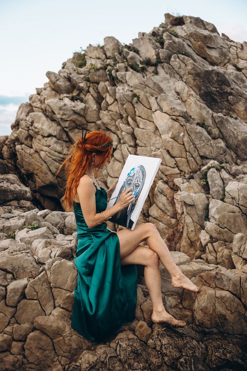 Free A woman wearing a green dress paints on a canvas while seated on rocks. Stock Photo