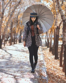 Stylish woman walks with umbrella amidst fall leaves on a sunny day.